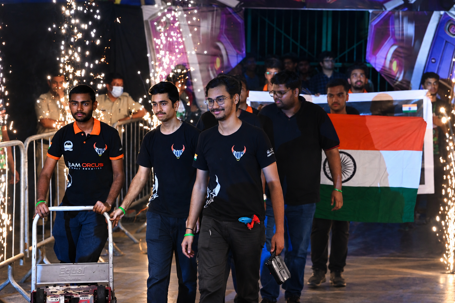 An Indian team member holding a robot controller in the arena, with teammates waving the Indian flag behind